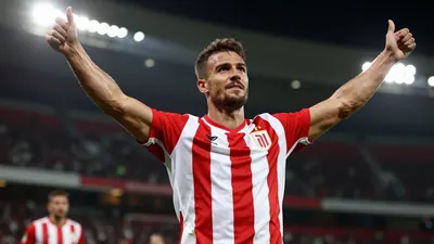 Photorealistic image of a muscular male football player in a red and white striped jersey, celebrating a goal with arms...