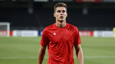 A young, athletic male striker in a solid red jersey, standing on a football pitch with a determined expression, ready to...