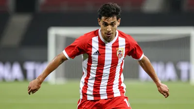 A tight action shot of a young, athletic male footballer in a red and white striped jersey, mid-dribble with the ball at...