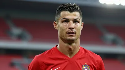 A 37-year-old experienced midfielder with a confident expression, wearing a solid red football kit, standing in a stadium...