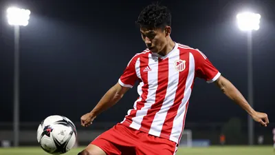 A young male football player in a red and white striped jersey, mid-kick with a football, in a training session under...