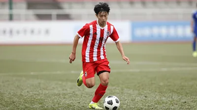 A tight action shot of a young Japanese midfielder in a red and white striped jersey, sprinting with the ball at his feet...