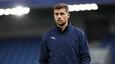A focused 25-30 year old male football coach in a solid dark blue training jacket, standing on the sidelines of a stadium