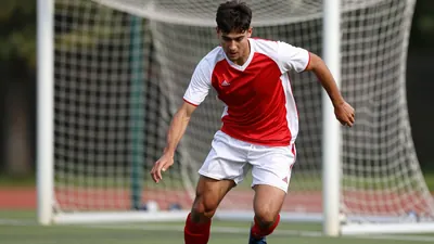 A young, muscular striker in a red and white jersey, head down in concentration, sprinting towards the goal with the ball...