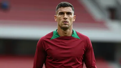 Mid-30s muscular forward, dark hair, intense eyes, wearing a plain dark-red and green training top (solid red base with...